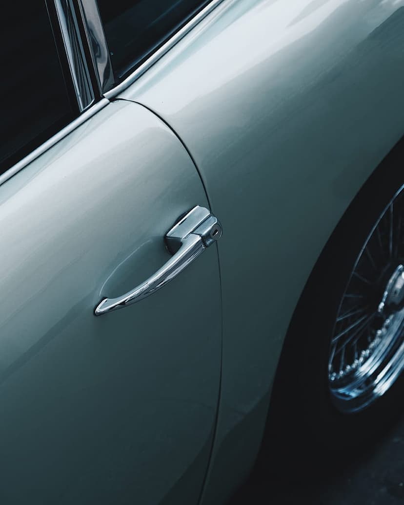 Chrome door handle and a wire-spoke wheel on a vintage car with muted bodywork and reflective metallic finishes.