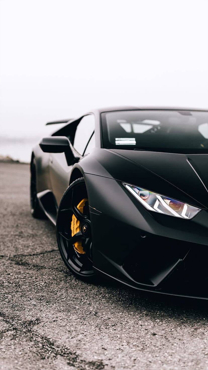 Matte black Lamborghini Huracan with yellow brake calipers sits on asphalt under a cloudy sky in a low-angle shot.