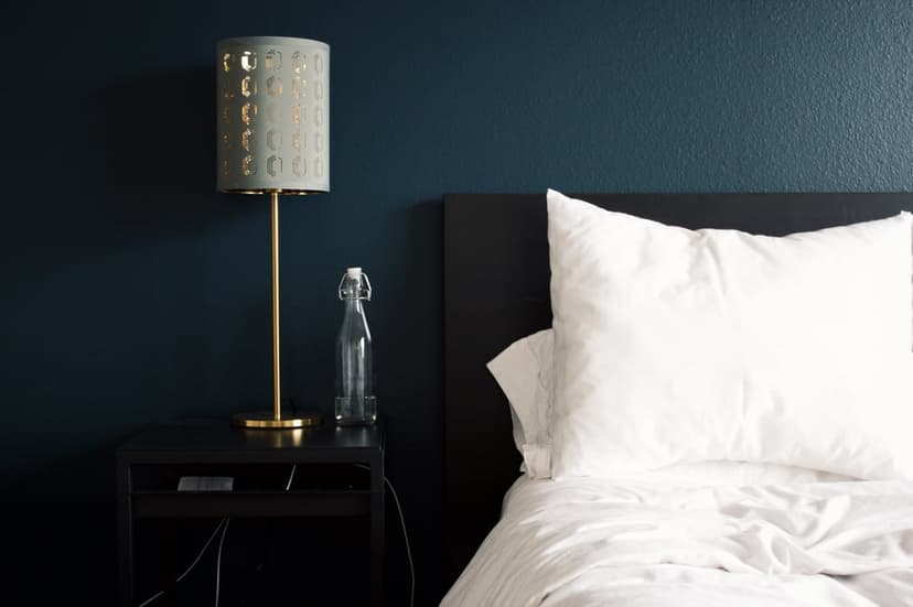 White bedding and plush pillows beside a minimalist black nightstand under a brass lamp against a navy blue wall.