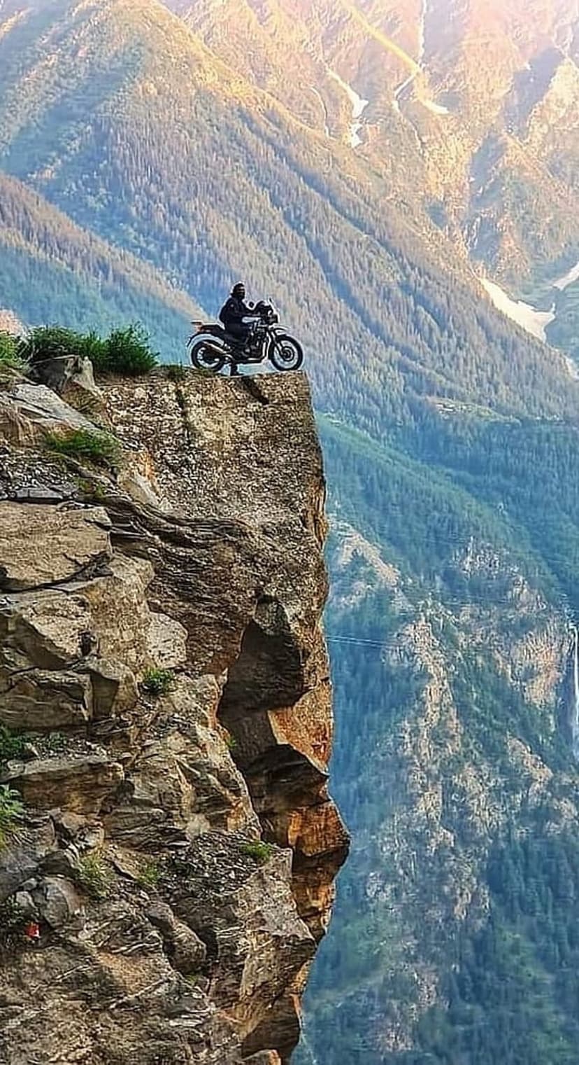 Motorcyclist sitting on a jagged rock ledge above a deep green valley with a distant waterfall under soft sunlight.