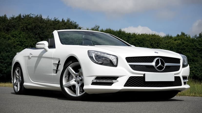 White Mercedes-Benz SL convertible features a low-angle view against green trees and a vibrant cloudy blue sky.