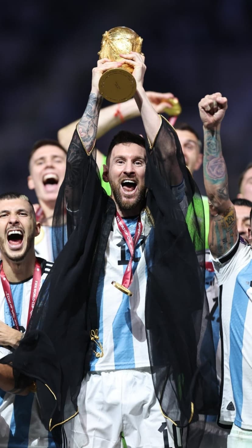 Lionel Messi lifting the golden World Cup trophy while wearing a black Bisht and Argentina national team jersey.