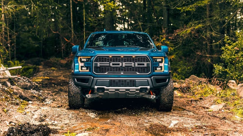 Front view of a teal Ford F-150 Raptor pickup truck on a muddy forest trail with glowing amber lights.