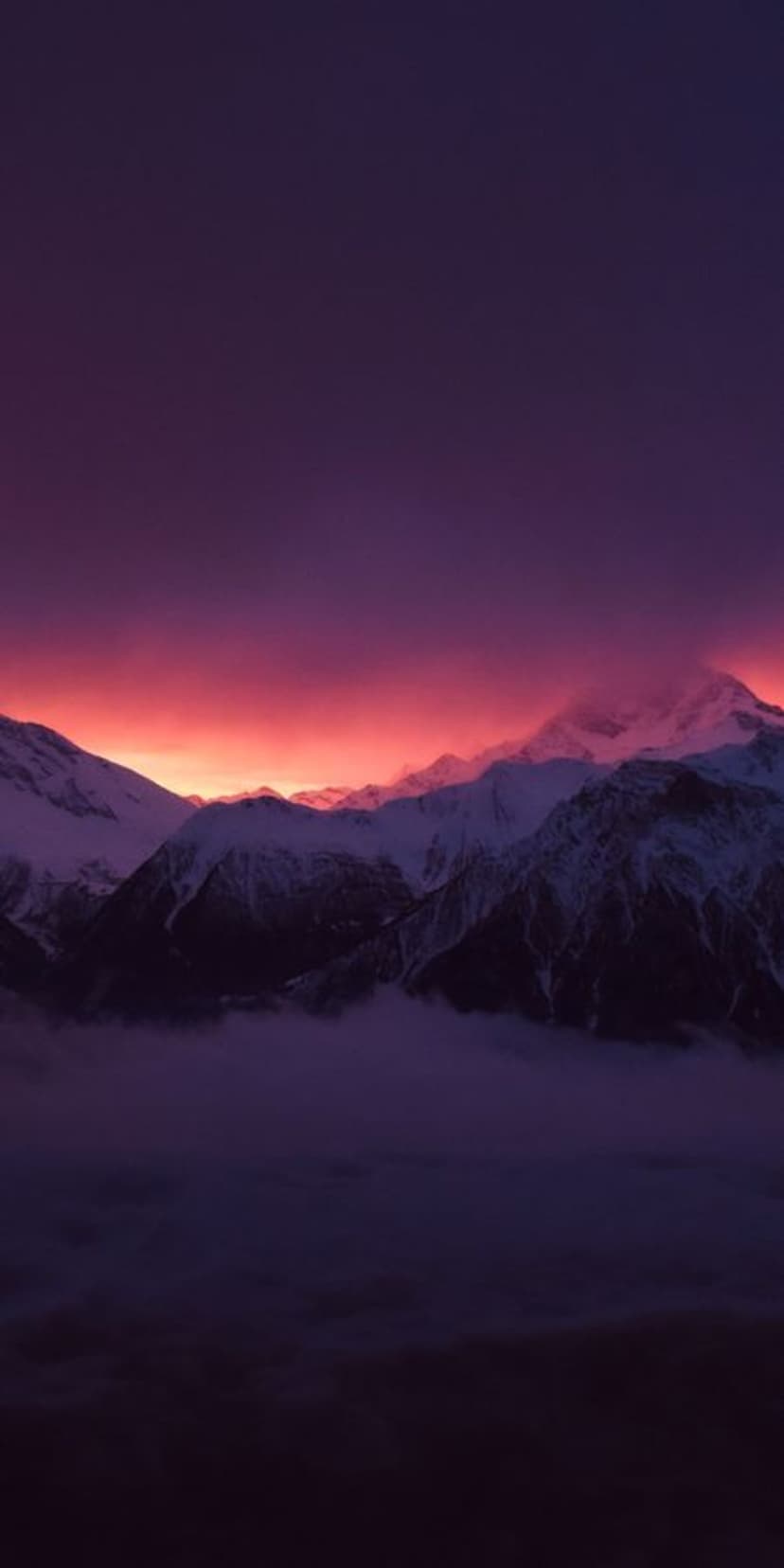 Snow-dusted mountain peaks glow red under a fiery sunset with clouds clinging to the rugged alpine slopes.