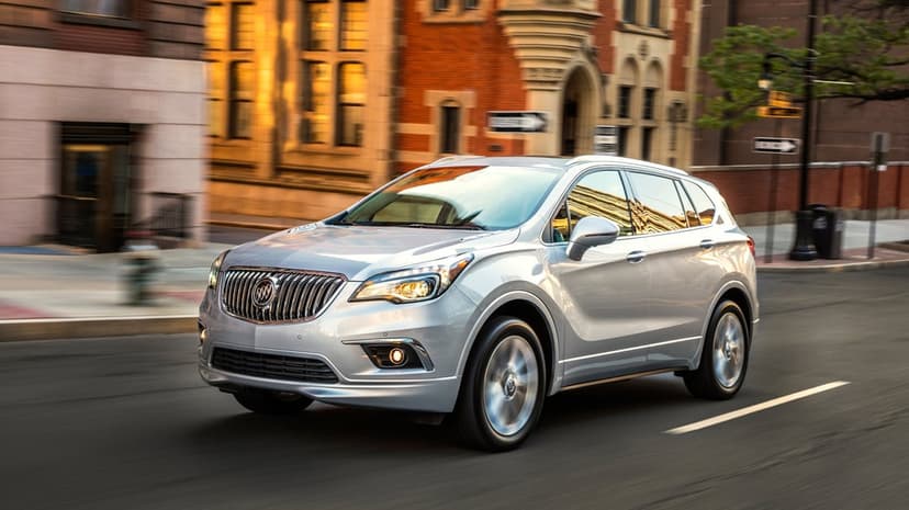 Silver Buick Envision SUV driving on a city street at sunset with golden light reflecting on its metallic body.