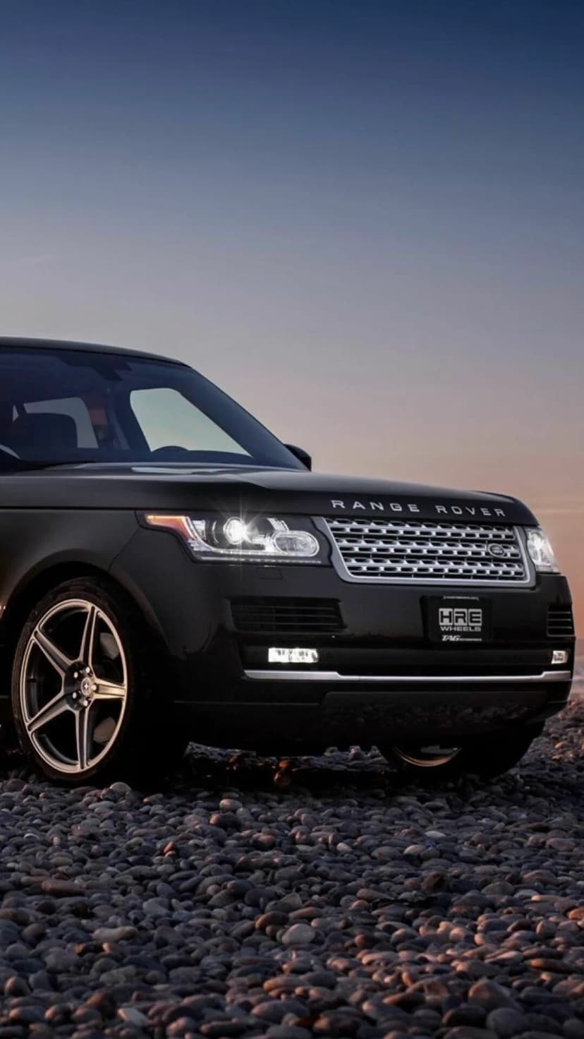 Matte black Range Rover parked on smooth beach pebbles with glowing headlights against a sunset sky.