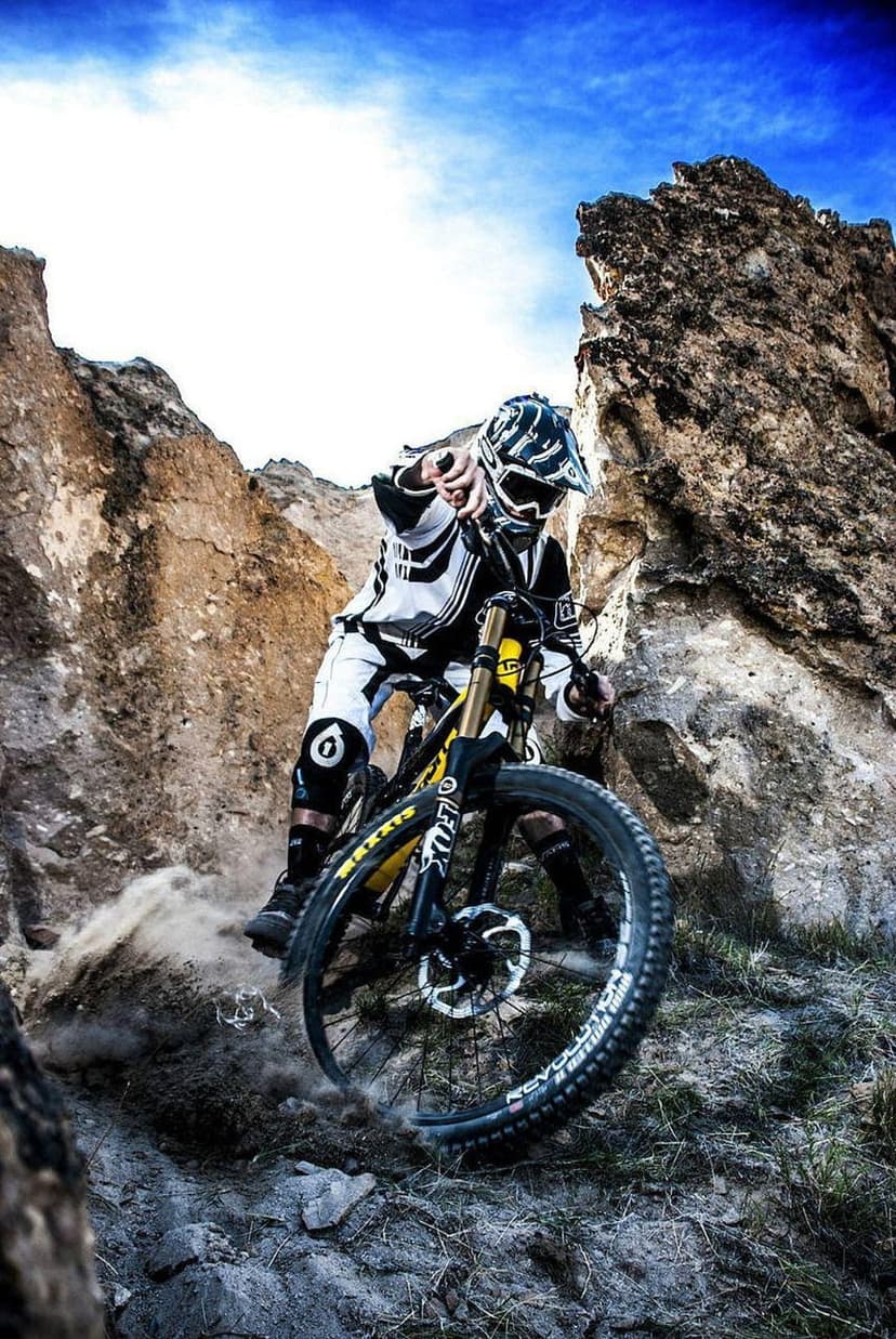 Mountain biker in full gear navigates a steep rocky ridge under a blue sky, kicking up clouds of dust mid-descent.