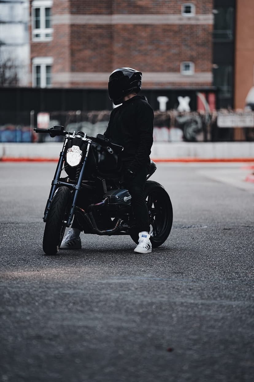 Person in black motorcycle gear and white sneakers sits on a dark bike against a blurred brick wall backdrop.