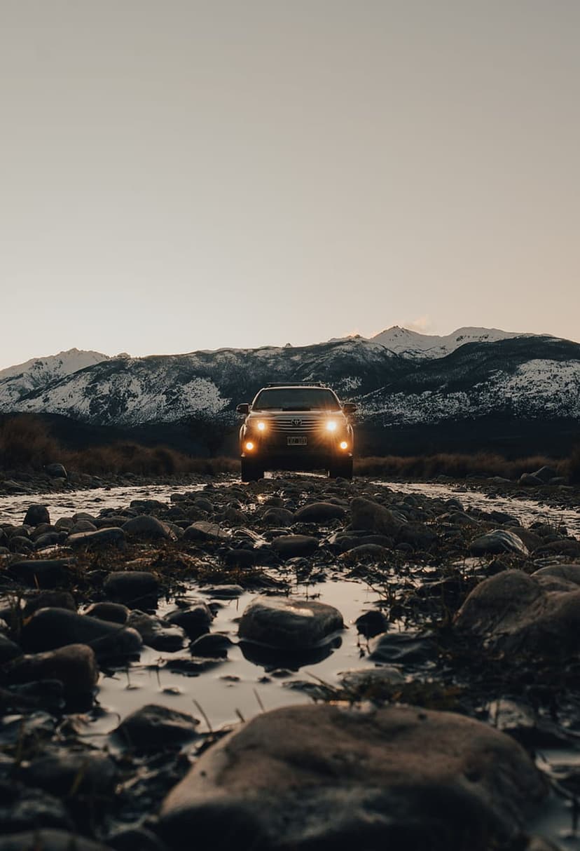 SUV with glowing headlights crosses a rocky river toward snowy mountain peaks under a dark, dusky twilight sky.
