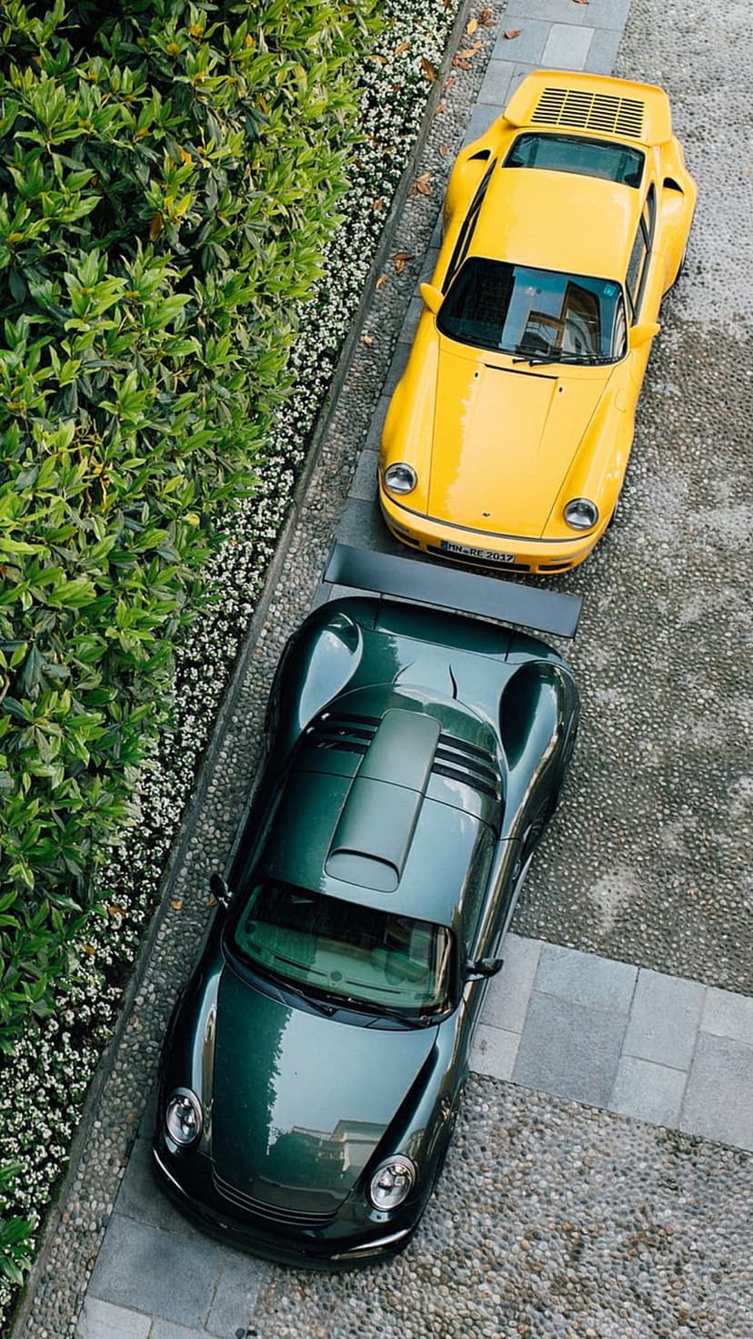 Yellow and dark green vintage Porsche cars parked on a gray cobblestone driveway surrounded by lush green leaves.
