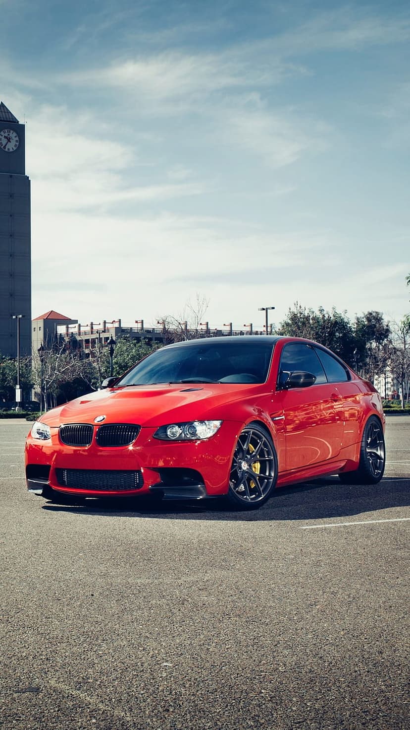 Red BMW M3 Coupe with yellow brake calipers parked in a lot with a clock tower under a cloudy sky.