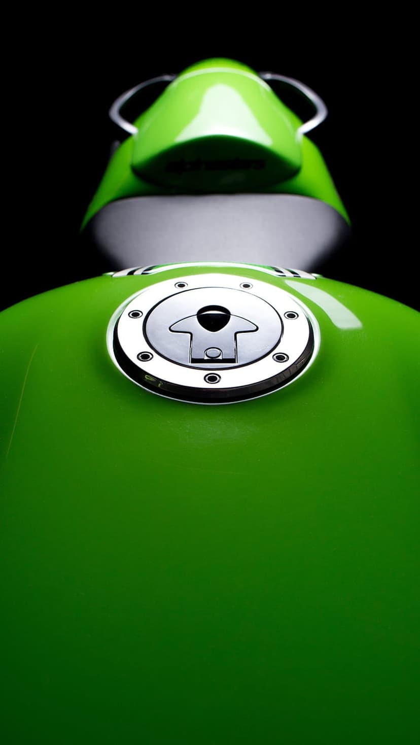 Green motorcycle fuel tank with a polished chrome cap and hex bolts isolated against a dark, shadowy background.