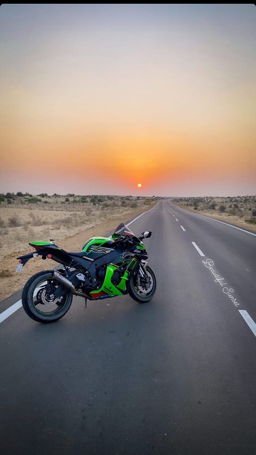 Green sportbike parked on a desert highway shoulder under a vibrant orange sunset with a distant horizon line.