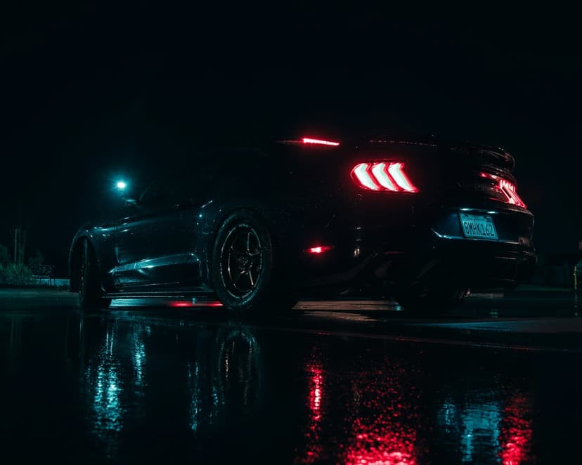 Ford Mustang GT rear view on a rain-slicked road at night with glowing red taillights and blue neon reflections.