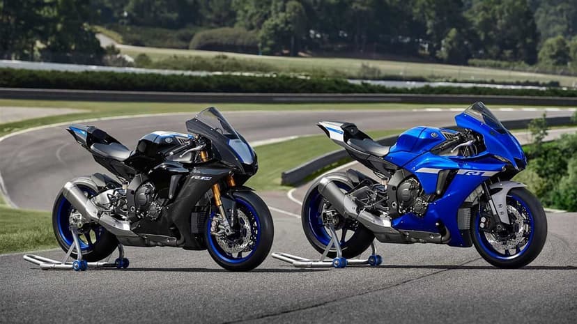 Black carbon fiber and blue Yamaha motorcycles parked on a racetrack under a clear sky with green hills.