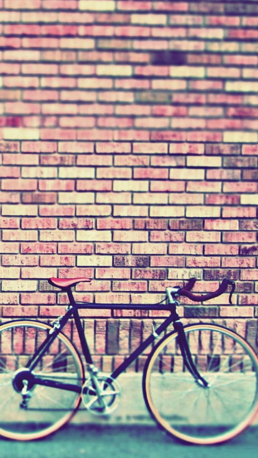Vintage black bicycle with a red leather seat leans against a weathered red brick wall in soft lighting.