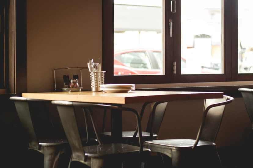 Wooden cafe table with stacked plates and metal chairs by a window overlooking a blurred red car on a city street.