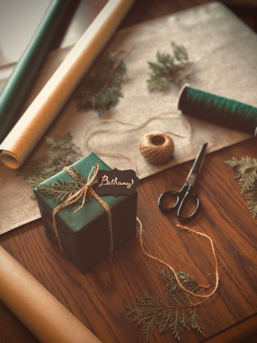 Dark green gift box with pine sprigs and Bethany tag on a wooden table surrounded by twine and scissors.