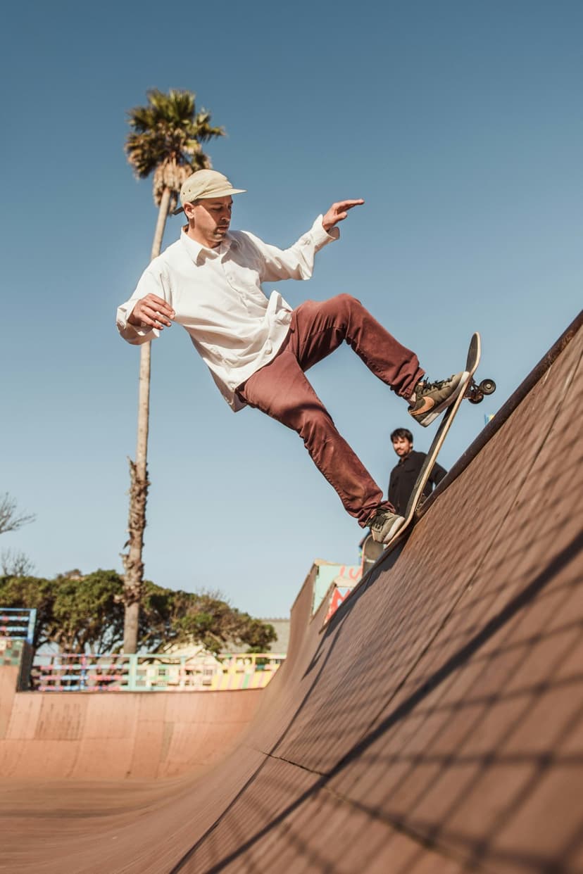 Skateboarder in a white shirt performs a mid-air trick on a wooden ramp under a clear sky with a palm tree.