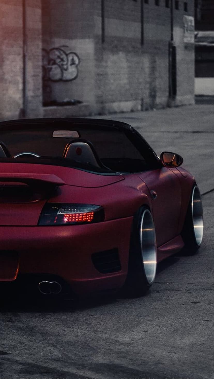 Red Porsche convertible rear view parked in a dark alley with graffiti on a brick wall under dim night lighting.