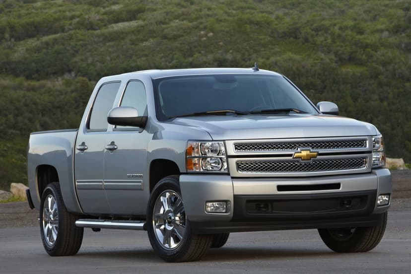 Silver Chevrolet Silverado pickup truck with chrome grille parked on asphalt against a forested hill background.