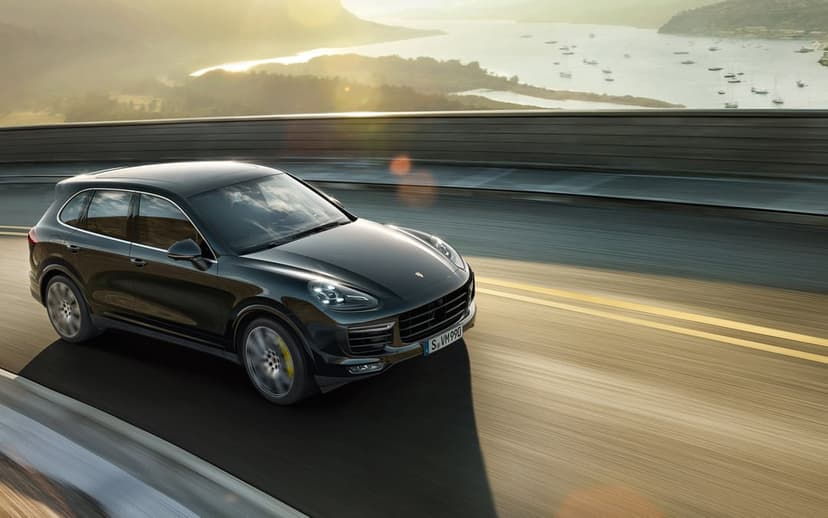 Black Porsche Cayenne driving fast on a coastal bridge at sunset with sailboats in a misty bay background.
