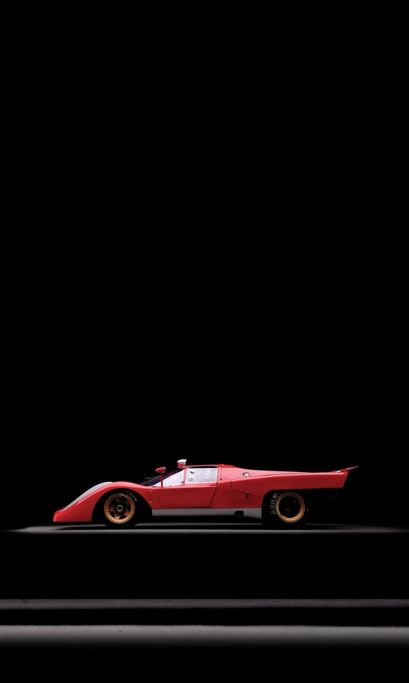 Classic red racing car with golden rims sits at a low angle against a pitch-black background in studio lighting.