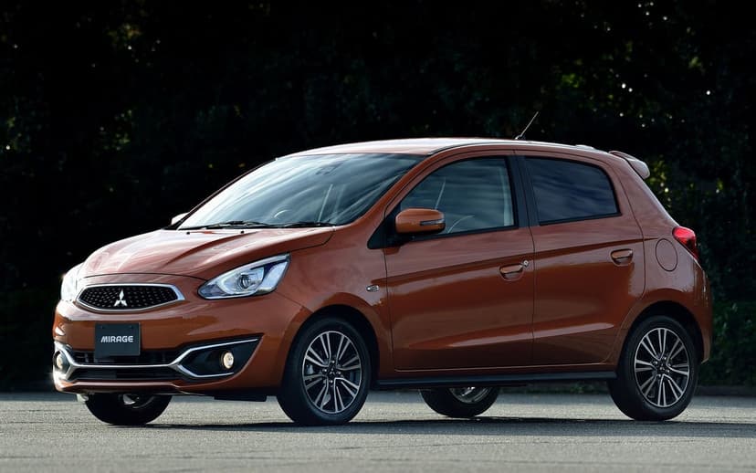 Orange Mitsubishi Mirage hatchback parked on asphalt against a dark blurred forest background in daylight.