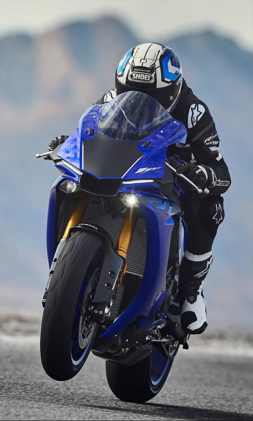 Rider on a blue Yamaha R1 motorcycle performing a wheelie on an asphalt road with blurred mountain background.