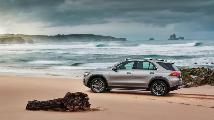 Silver Mercedes-Benz SUV on a sandy beach with crashing ocean waves, coastal cliffs, and driftwood log.