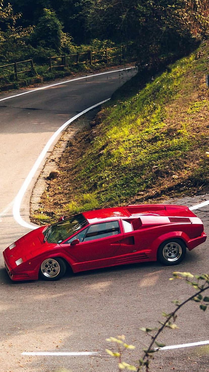 Red Lamborghini Countach supercar drives along a winding asphalt road through rolling green hills in sunshine.