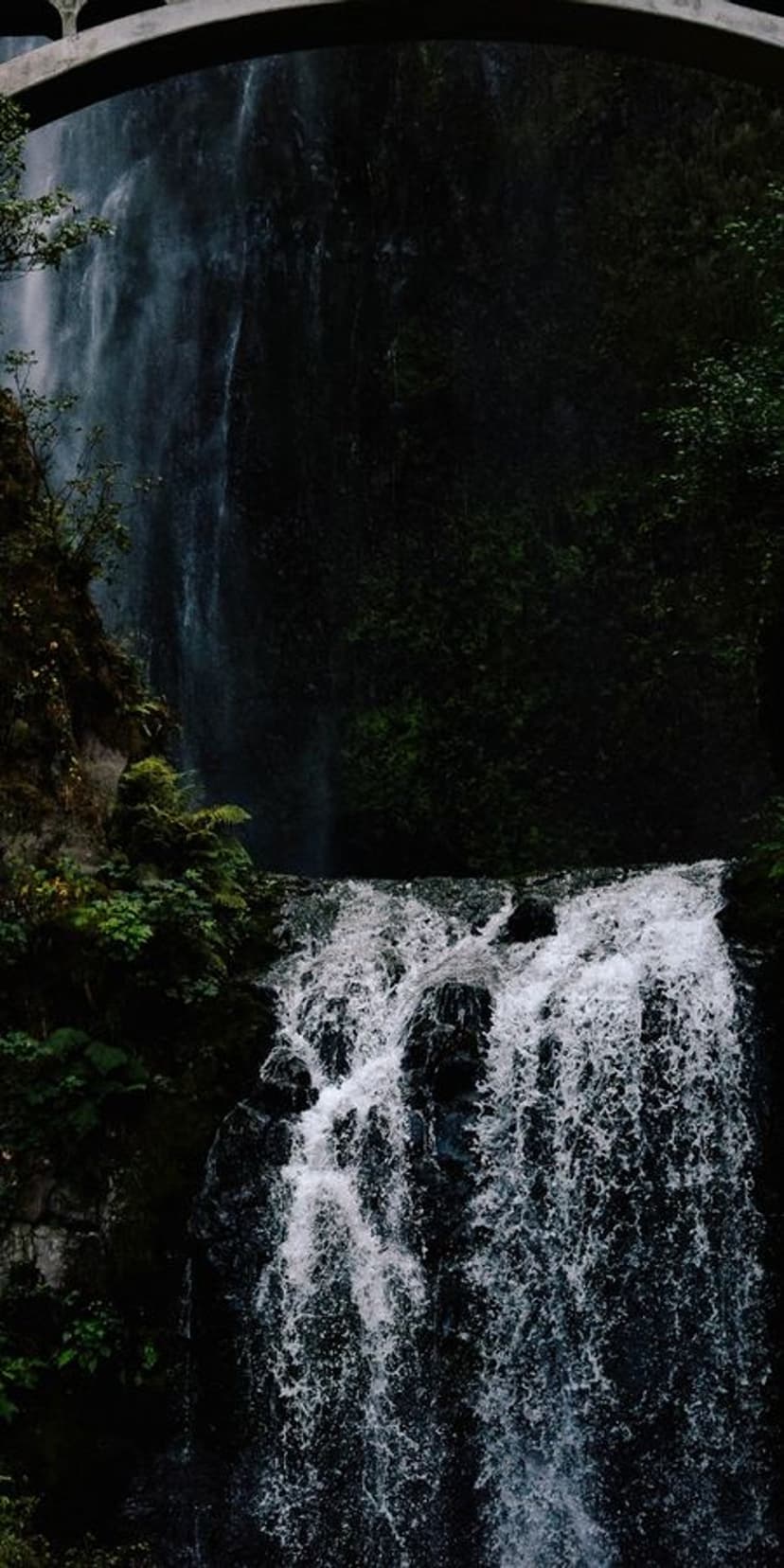 Tiered white waterfall rushing over mossy black rocks beneath a stone arch bridge in a lush forest setting.