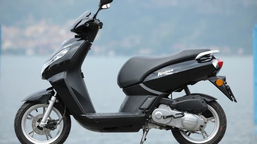 Profile of a black Peugeot Kisbee scooter on a kickstand with a blurred blue-grey water background.