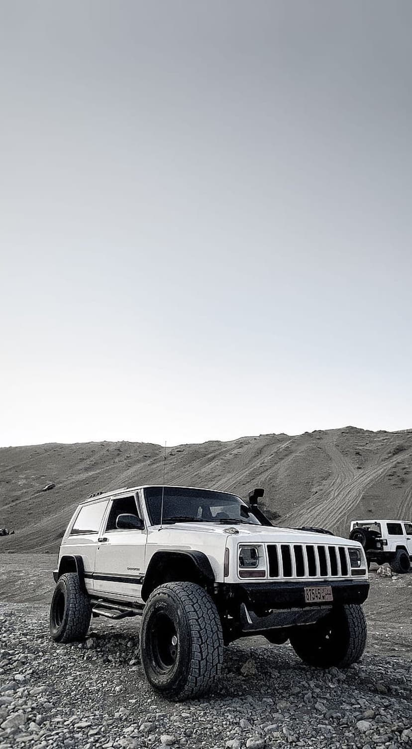 White two-door Jeep Cherokee with oversized tires parked on gravel terrain with sandy hills in the background.