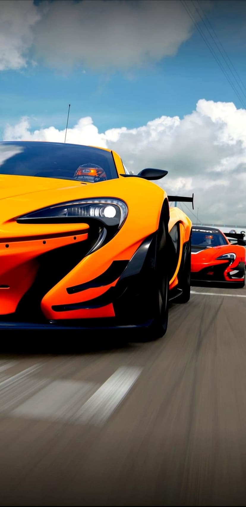 Two McLaren race cars in orange and red liveries compete on a sunlit asphalt track under a blue sky with clouds.
