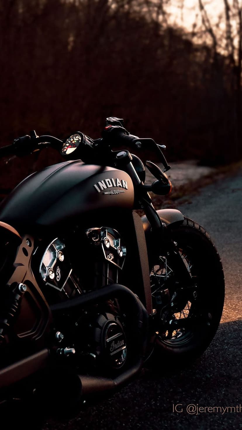 Matte black Indian Scout motorcycle parked on a forest road during sunset with chrome engine details visible.