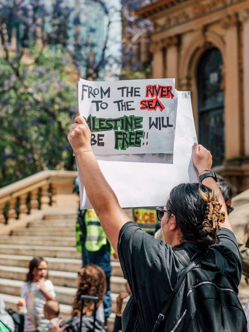 Protester holds a handwritten sign with red and green text during a sunny outdoor political rally on stone steps.