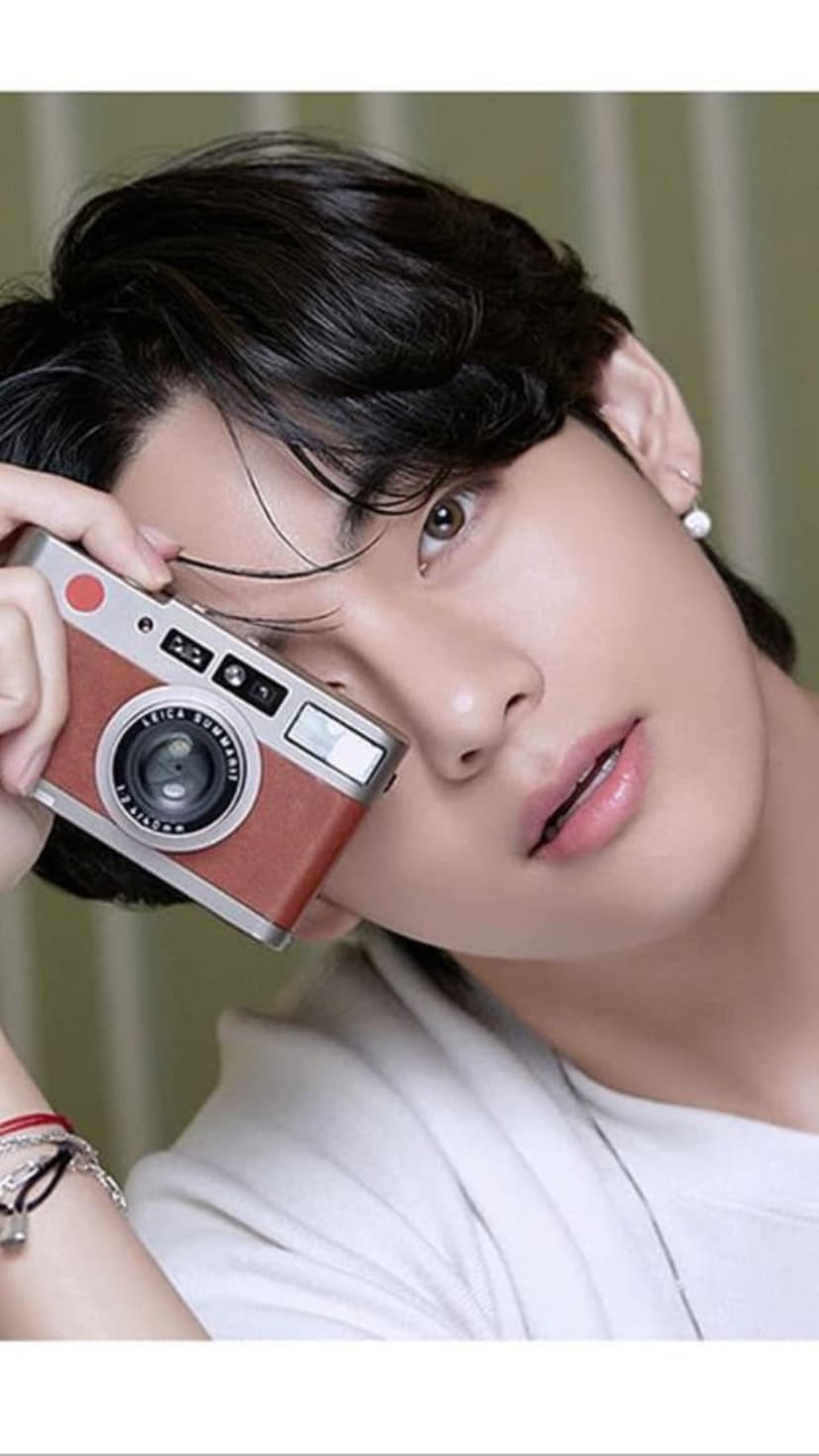 Kim Taehyung holds a vintage Leica camera to his face against a blurred green background in a vertical portrait.
