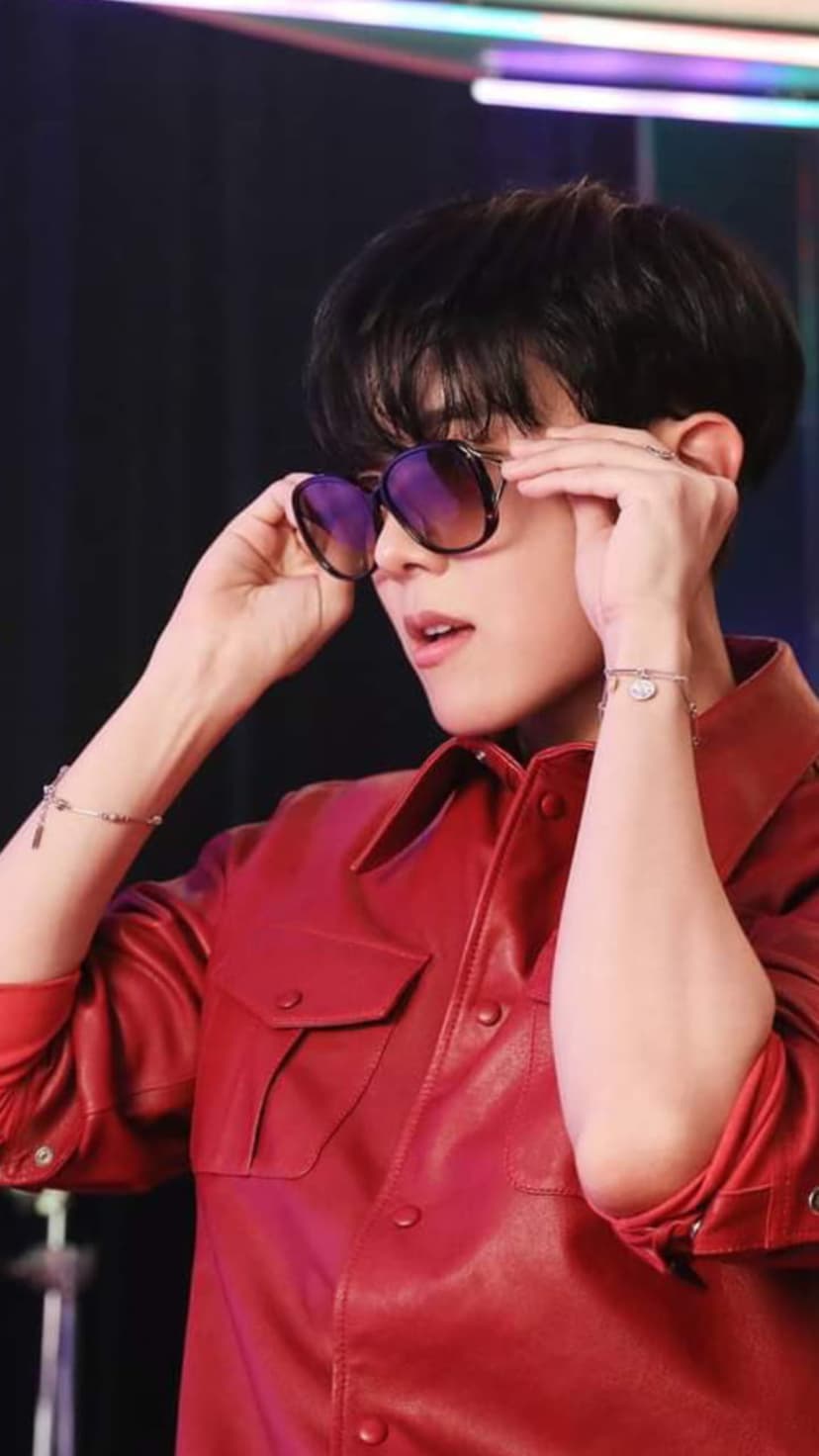 Man in a red leather jacket and dark sunglasses adjusts his eyewear against a blurry neon light background.