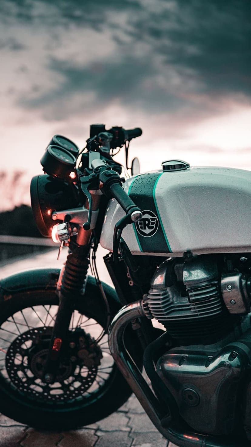 Royal Enfield motorcycle with rain droplets on its white fuel tank parked under a moody, overcast twilight sky.