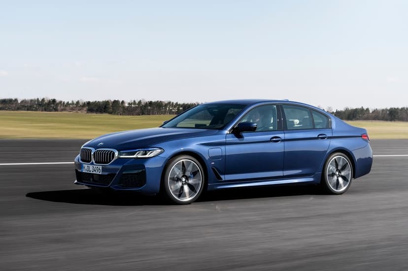 Blue BMW sedan driving on a track with motion-blurred wheels against a background of green fields and clear sky.