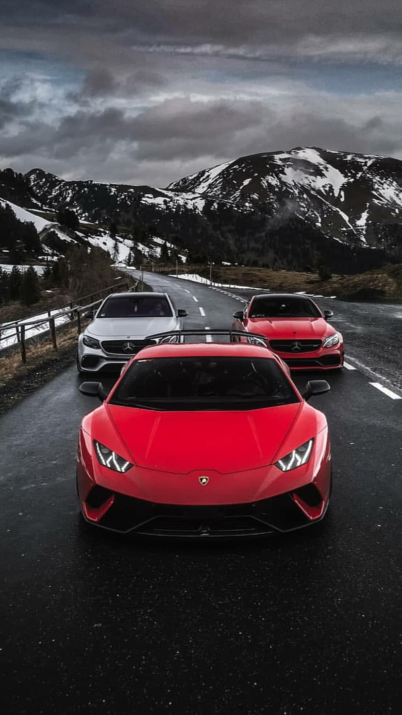 Red Lamborghini and Mercedes cars drive on a wet asphalt road through a snowy alpine mountain landscape.
