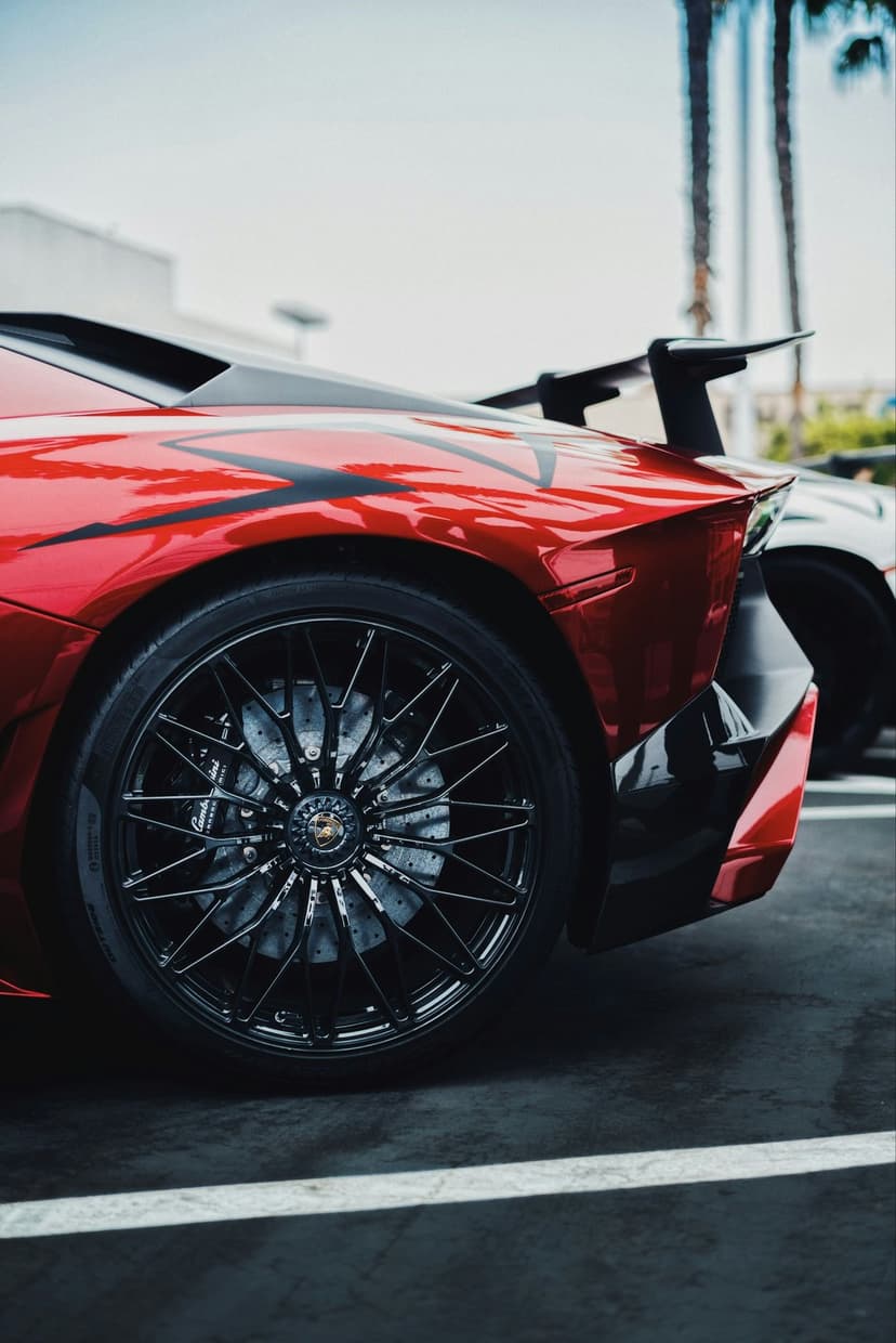 Red Lamborghini Aventador rear view with a large black spoiler and multi-spoke rims against blurred palm trees.