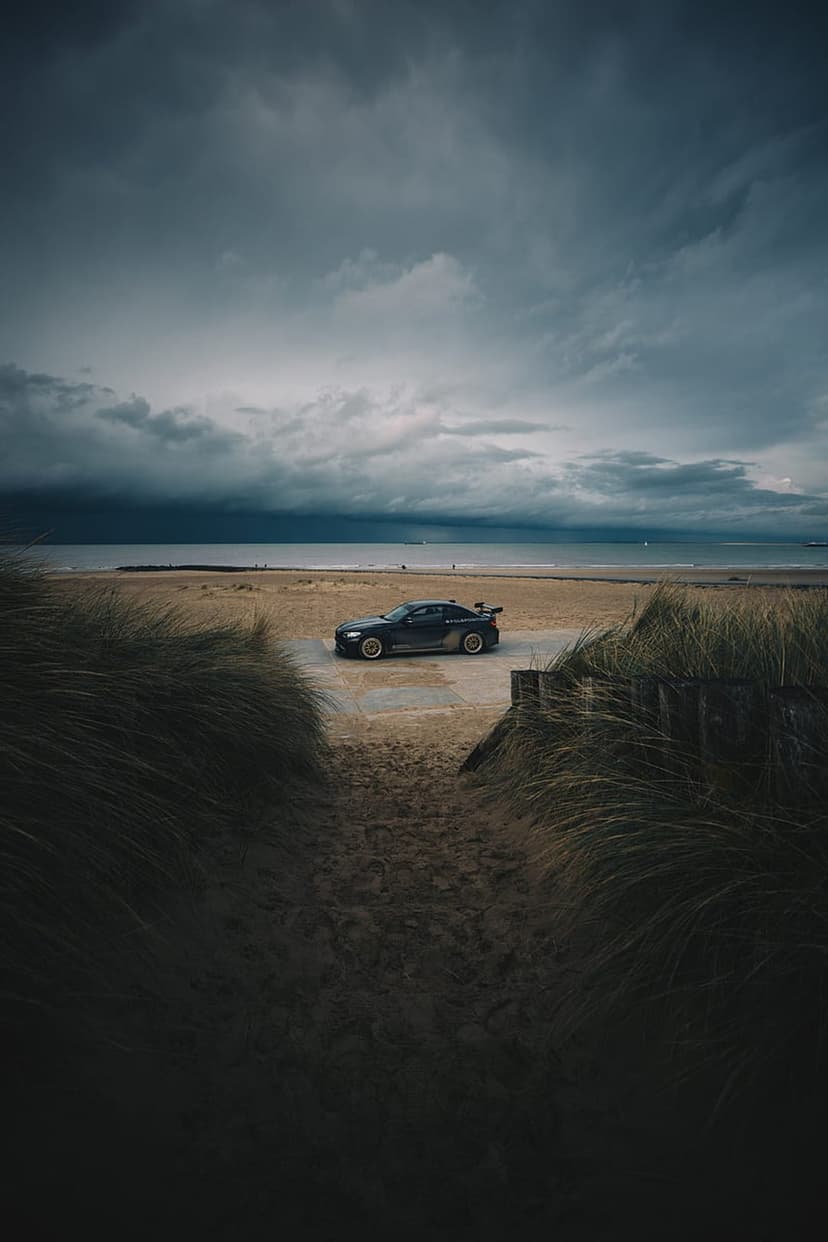 Black sports car parked on a sandy beach with tall grass in the foreground under dark, turbulent storm clouds.