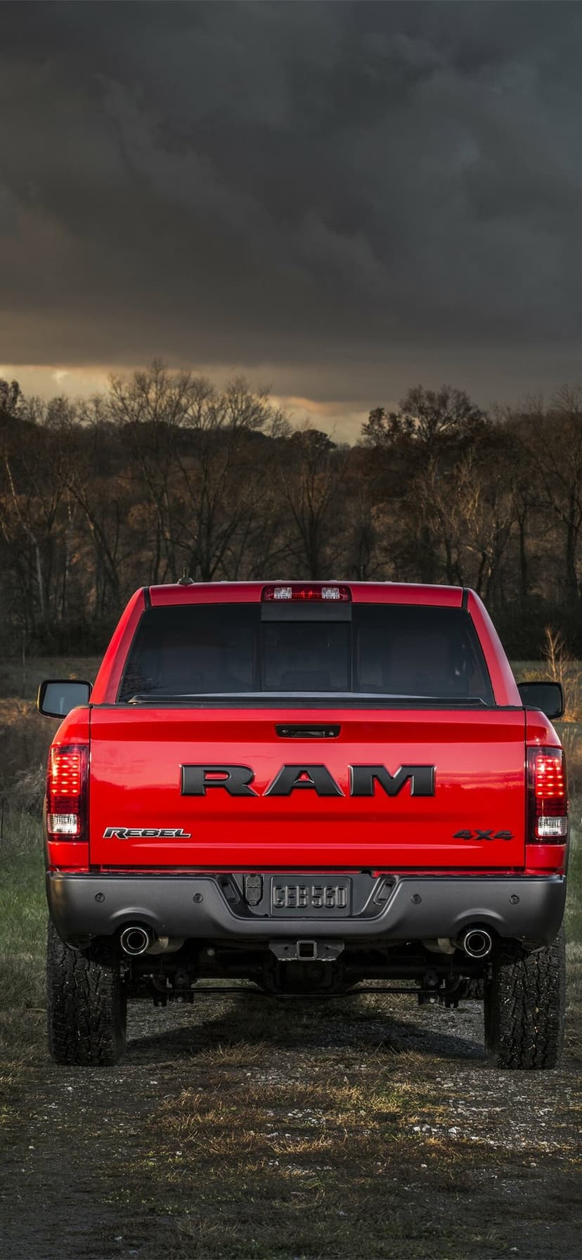 Rear view of a red Ram Rebel truck with black accents and dual exhaust under dark clouds and bare winter trees.