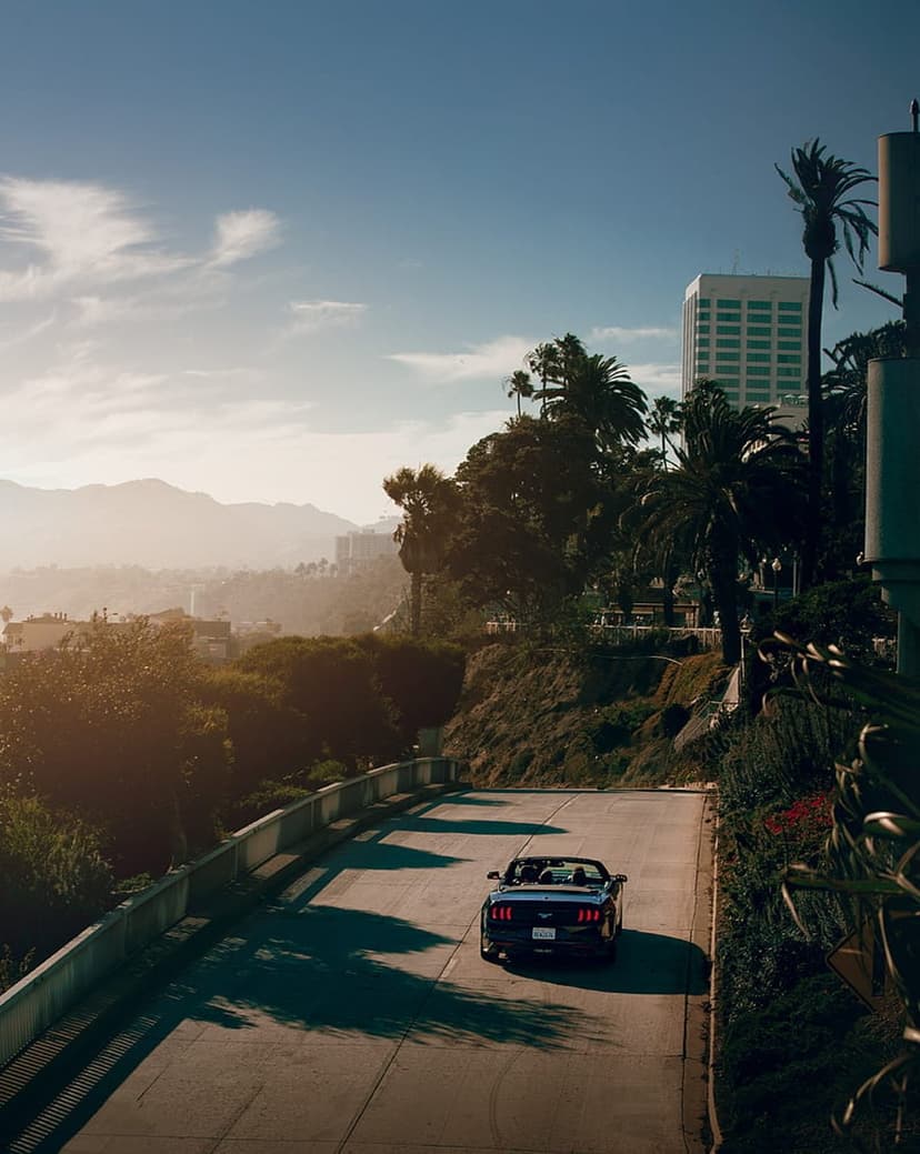 Black convertible Mustang drives on a curved road with mountains and a city skyline in the hazy distance.