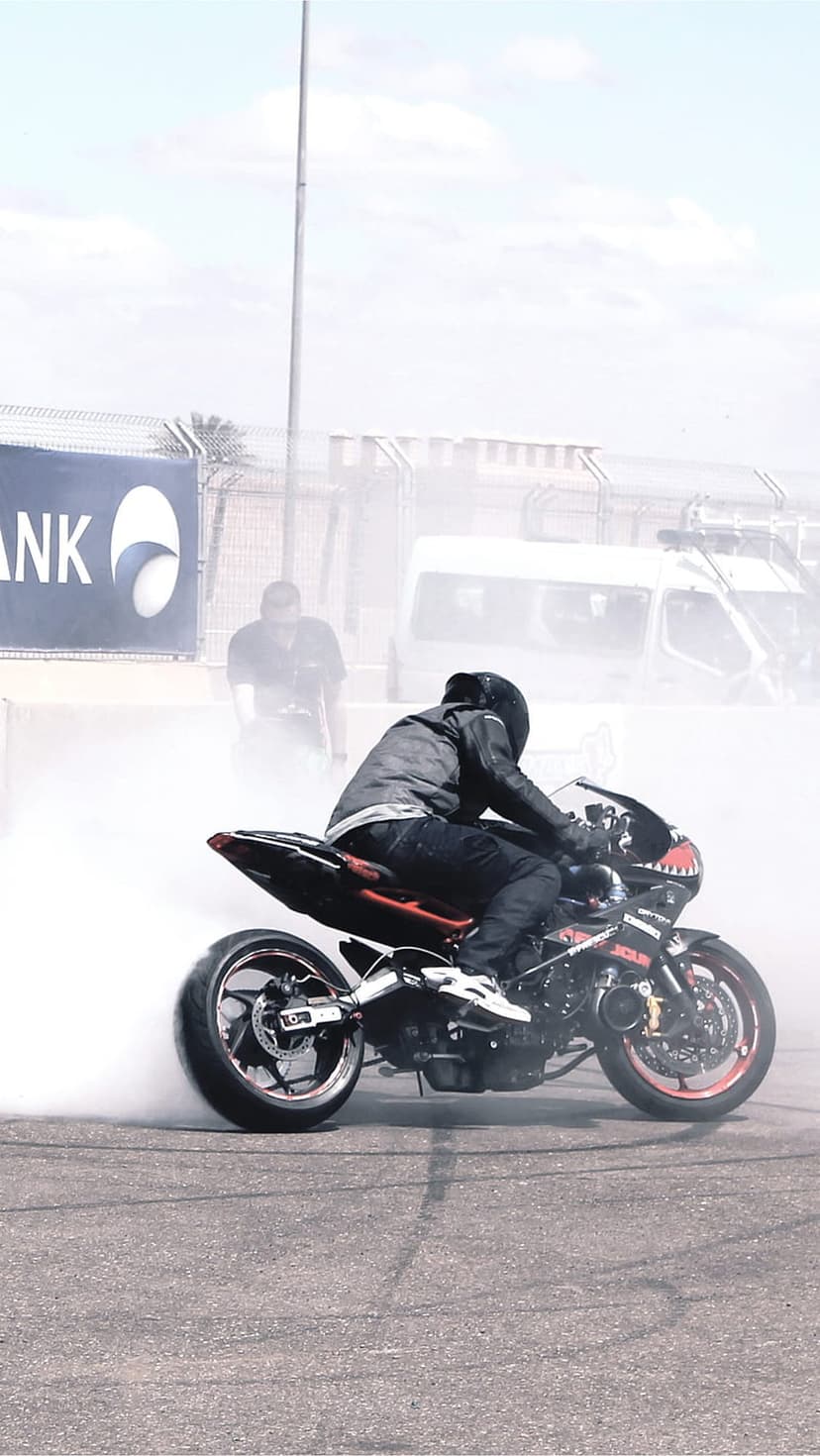 Motorcyclist in full gear leaning over a sportbike while creating a massive cloud of white smoke from a burnout.