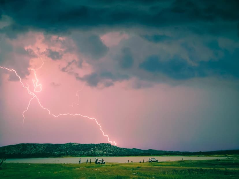 Pink lightning bolt illuminates a dark lake and hills while people watch from a shoreline with a car.