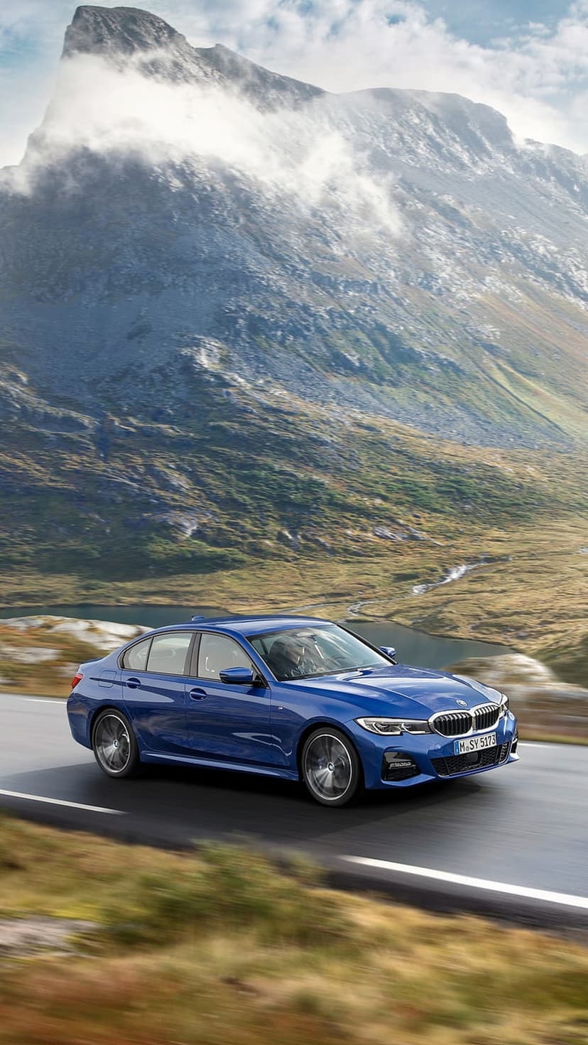 Blue BMW sedan driving on a winding road through jagged mountains under a cloudy sky in portrait view.