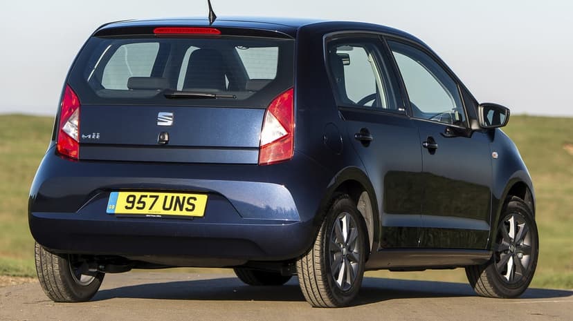 Dark blue SEAT Mii hatchback parked on a quiet road with a blurred green field and clear sky in the background.